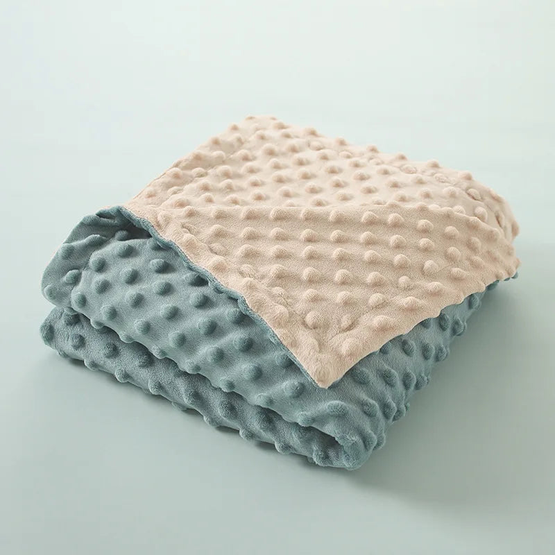 Couverture bébé réversible pliée montrant  deux faces - bleu-vert et beige - avec texture à motifs en relief démontrant  polyvalence du produit