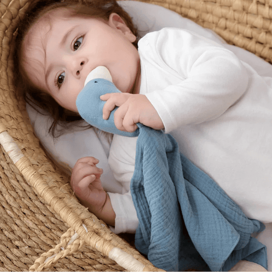 Bébé réconforté dans un couffin, mâchant son Doudou Bebe canard en mousseline bleue